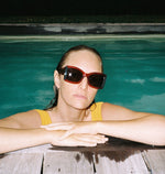A girl wearing red framed sunglasses with dark tinted lenses by the pool. 