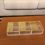 A three-compartment acrylic organizer box with gold-colored geometric patterns on the lids, placed on a wooden table with a sofa in the background.