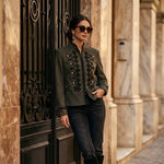 Model wearing KIDDING olive green suede military Napoleon jacket with black braided scroll frogging, gold dome buttons, Mandarin collar, and black piping paired with dark jeans and boots