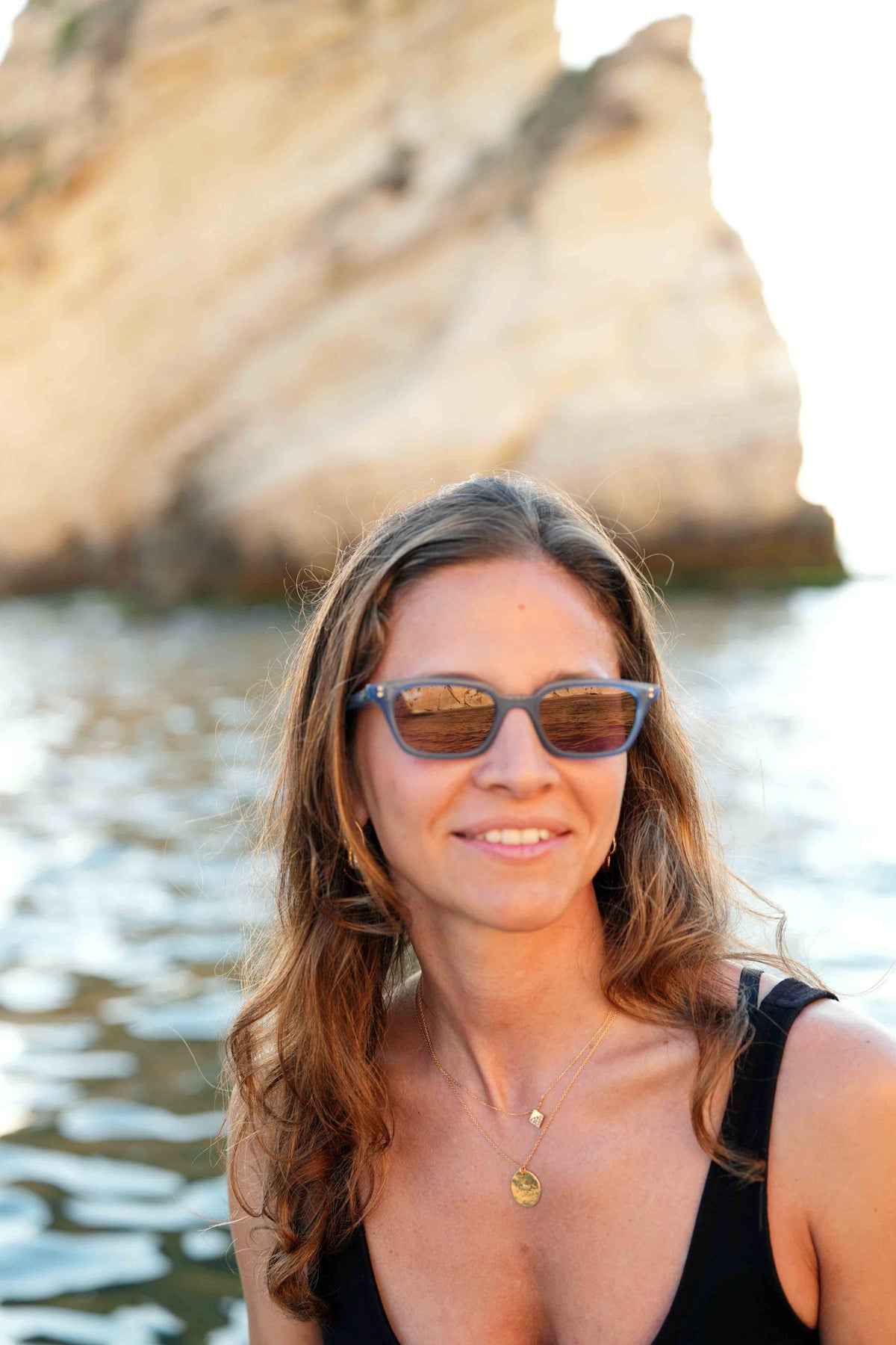 A woman with long brown hair wearing dark sunglasses and a black top, standing outdoors in front of water and a large sandy rock formation.