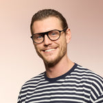 A man wearing black framed eyeglasses and black striped shirt on a pink background. 