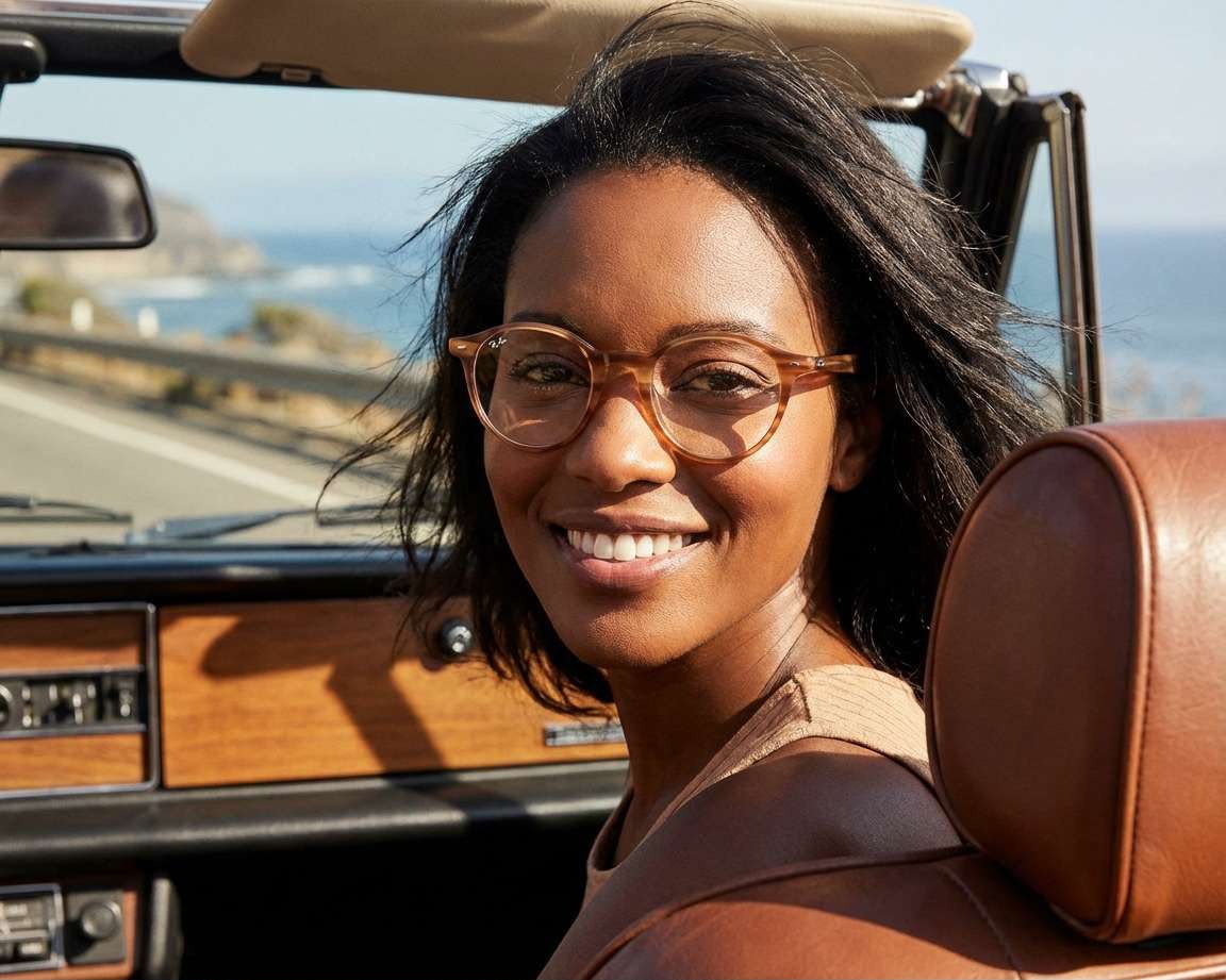 A woman wearing round eyeglasses and looking at the camera, with a pretty view behind her. 