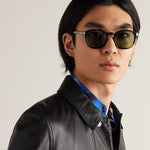 A close-up portrait of a man wearing black round-frame sunglasses with dark lenses, a black leather jacket, and a blue patterned shirt collar, set against a plain light gray background.
