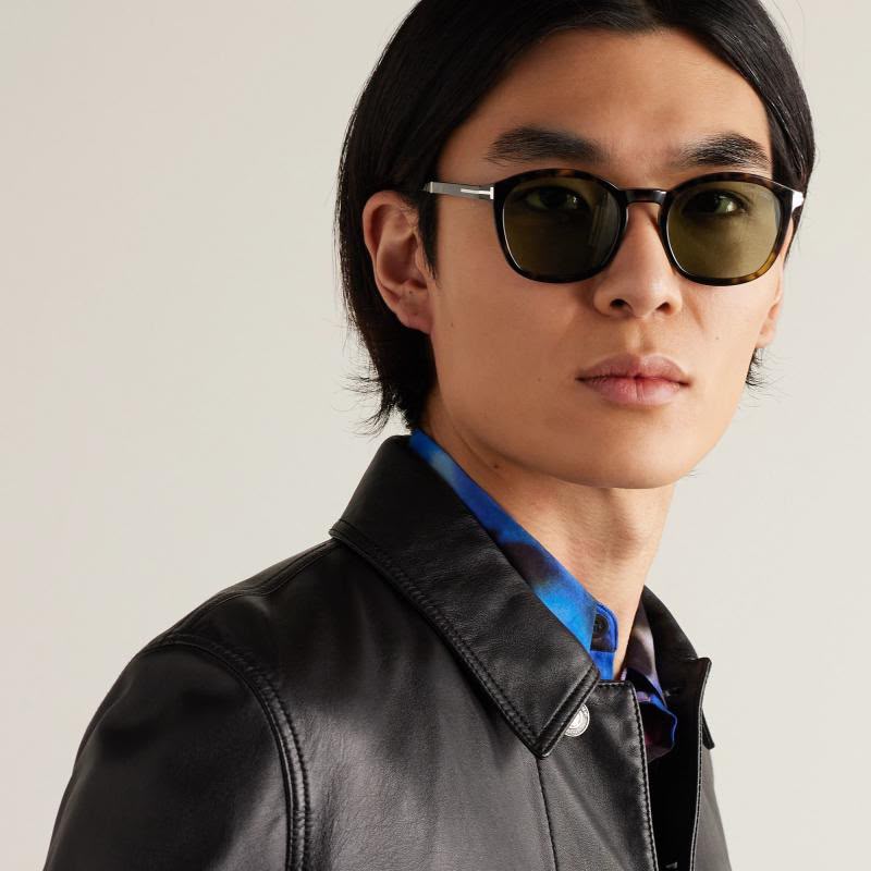 A close-up portrait of a man wearing black round-frame sunglasses with dark lenses, a black leather jacket, and a blue patterned shirt collar, set against a plain light gray background.