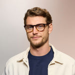 A man wearing tortoiseshell framed eyeglasses on a light purple background. 