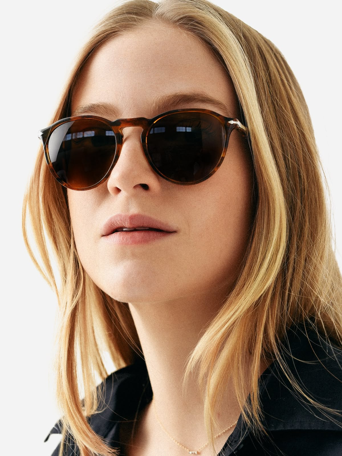A close-up of a woman with blonde hair wearing round tortoiseshell sunglasses with dark brown lenses and silver metal frames, set against a clean white background.