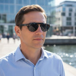 A mean wearing tortoiseshell framed sunglasses with dark tinted lenses and a blue shirt. 