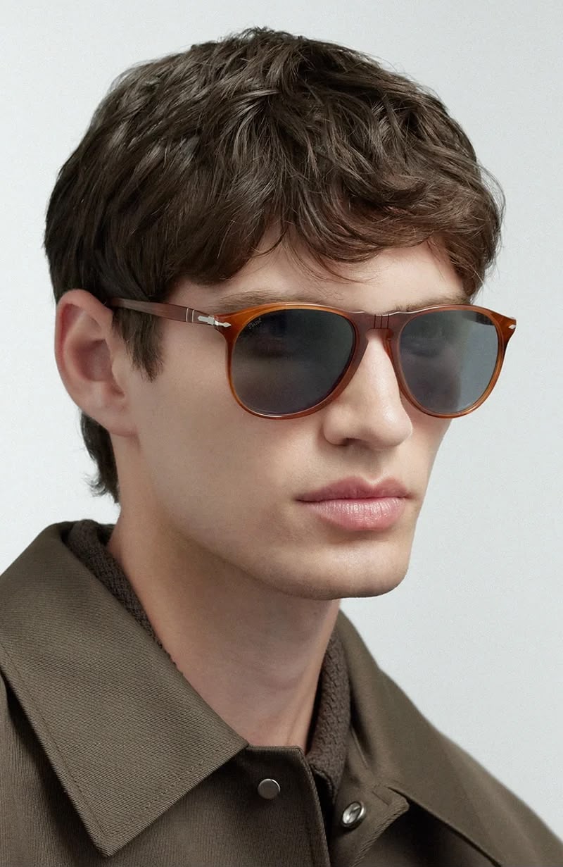 A man wearing tortoiseshell framed sunglasses with dark lenses on a white background. 