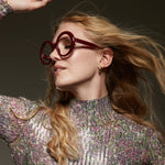 Side profile of a girl wearing burgundy framed eyeglasses and a shining top on a dark background. 