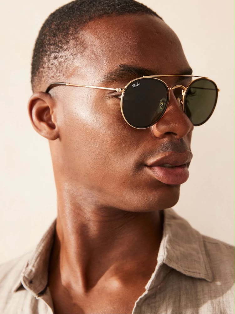 A close-up of a man wearing Ray-Ban aviator sunglasses with gold frames and dark green lenses, paired with a light beige linen shirt against a warm beige background.
