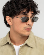 A close-up of a man wearing slim, geometric square-frame sunglasses with dark lenses and thin gold-toned metal arms, paired with a beige corduroy shirt over a white t-shirt, against a light gray background.