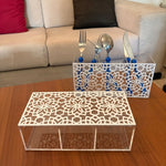 Two white laser-cut acrylic organizer trays with floral patterns, one holding silver cutlery in blue holders, displayed on a wooden table in front of a beige sofa.