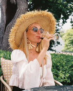 A woman outdoors wearing a large straw sun hat, sunglasses, and a white blouse, holding a glass of red wine. She is seated in a chair with green foliage and a tree trunk in the background.