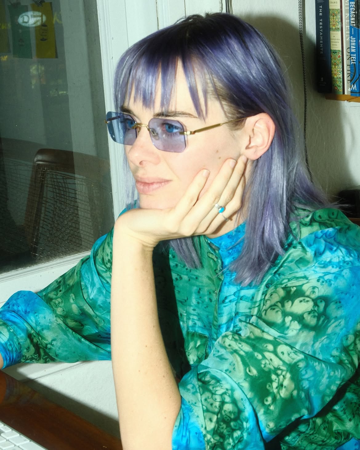 A woman with purple hair and sunglasses rests her chin on her hand, wearing a green and blue patterned blouse in an indoor setting.
